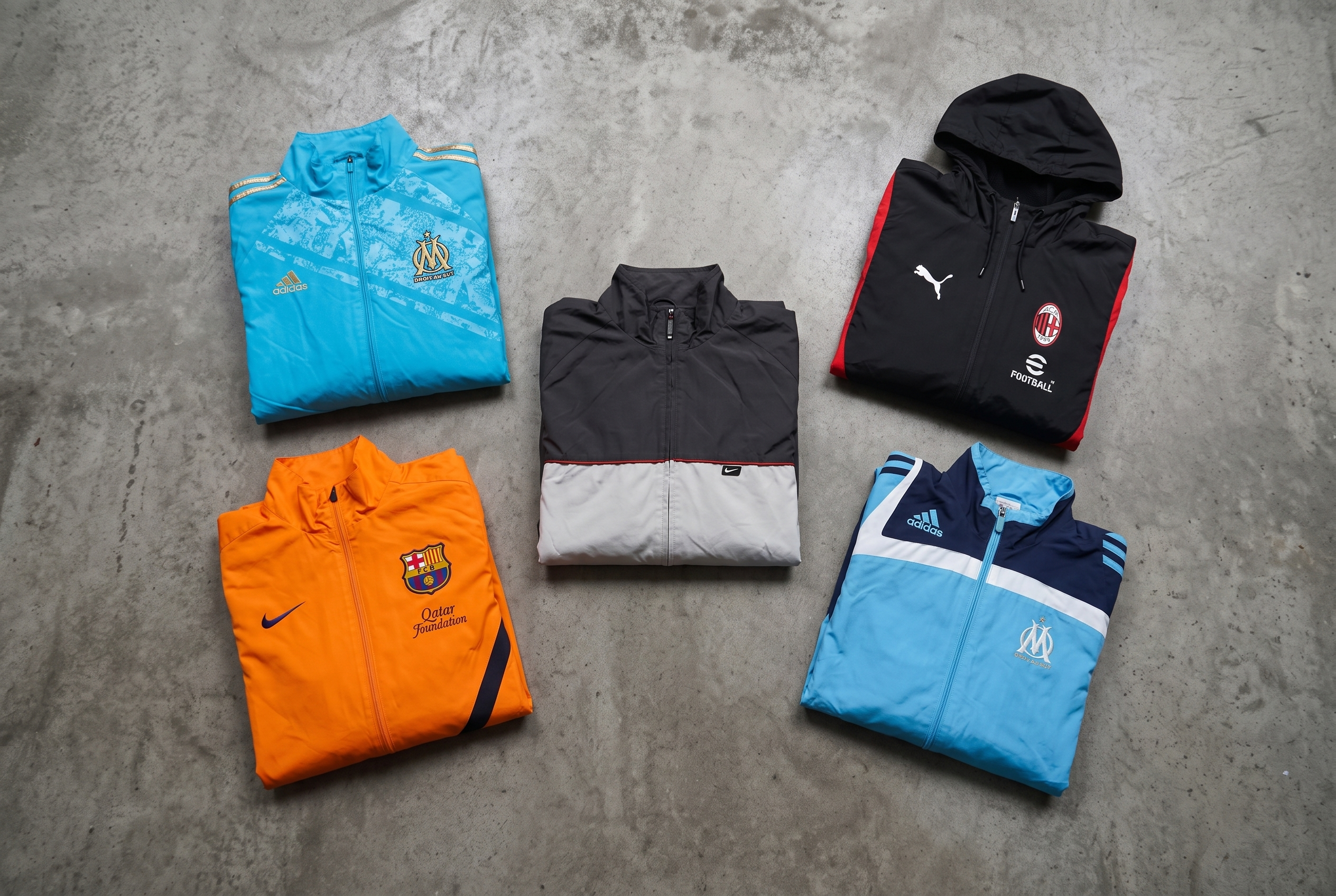 Fünf zusammengelegte Vintage-Sportjacken in Blau, Orange, Grau und Schwarz auf Betonboden